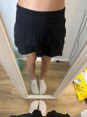 Svart kortkjol från Gina Tricot - Svart kortkjol från Gina Tricot med bred smock i midjan och volangig nederdel. Kjolen har en enkel och stilren look som passar till många olika outfits. Tillverkad i ett lätt och mjukt material som känns bekvämt.