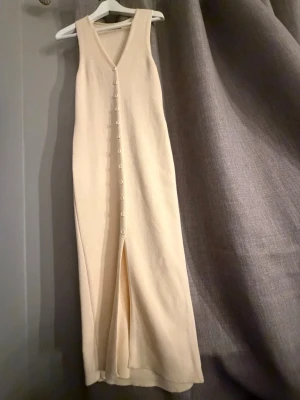 Helt ny Beige stickad klänning Gina Tricot - Helt ny Lång beige stickad klänning från Gina Tricot, den är ny med inga fläckar eller något tecken på andvändning. Den köptes för 500kr. Den har v-ringning och knappar hela vägen framtill. Klänningen är ärmlös och har en slits nedtill för extra rörelsefrihet. Perfekt för dig som gillar en enkel men stilren look.