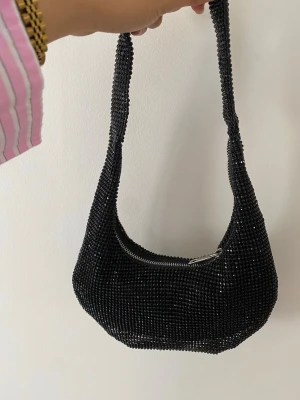 Svart axelväska med strassdetaljer från bik bok - Trendig svart handväska med glittrande strassdetaljer och en mjuk, rundad form. Väskan har ett kort axelband och stängs med en silverfärgad dragkedja. Perfekt accessoar för att lyfta vilken outfit som helst.