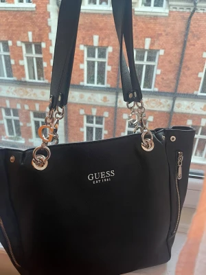 Svart handväska från Guess - Snygg svart handväska från Guess med silverfärgade detaljer och kedjor på handtagen. Väskan har Guess-logga framtill, dragkedjor på sidorna och ett stort G-hänge. Tillverkad i konstläder med struktur. Perfekt för dig som gillar stilrena accessoarer.