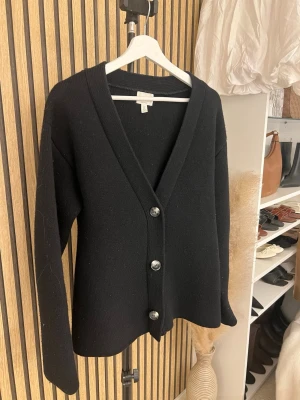 Svart ull kofta från H&M med knappar - Klassisk svart kofta från H&M med v-ringning och stora silvriga knappar framtill. Koftan är långärmad och har en enkel, stilren design som passar till det mesta. Perfekt att slänga över en t-shirt eller topp för en avslappnad look. Köpt för original priset.