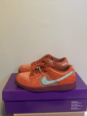 Nike SB Dunk Low Orange Label - Nike SB Dunk Low sneakers i orange mocka med ljusblå Swoosh och detaljer. Skorna har klassisk låg silhuett, vadderad plös och tåbox med perforeringar. Perfekta för dig som gillar streetwear och unika färgkombos.