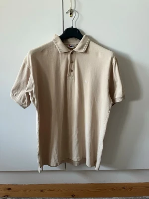 Beige pikétröja från Intersport strl L - Klassisk beige pikétröja från Intersport med krage och tre knappar framtill. Tillverkad i 100% bomull. Storlek L. Har lite missfärgningar på ryggen, se sista bild.