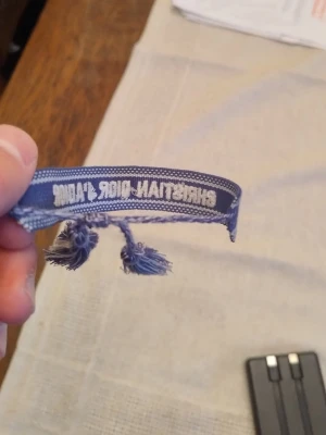 Blått armband Christian Dior J'Adior - Snyggt blått tygarmband från Christian Dior med texten 'J'Adior' broderad i vitt. Armbandet har justerbar knytning med tofsar i ändarna och är perfekt för att ge din outfit en exklusiv touch.