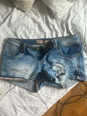 Blå slitna jeansshorts med nitar - Säljer ett par blå jeansshorts från monday pretty med slitningar och hål på framsidan, råa kanter och dekorativa nitar vid fickorna. Klassisk femficksmodell med dragkedja och knapp. Perfekta för en avslappnad och trendig look. Helt oanvända! Tyvärr för små för mig