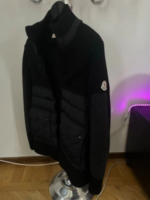 Svart cardiganjacka från Moncler - Snygg svart cardiganjacka från Moncler med stickade ärmar och quiltade paneler framtill. Jackan har hög krage, dragkedja och två stora fickor med lock. Moncler-logga på ärmen ger en exklusiv touch. Perfekt för dig som vill ha både stil och komfort.