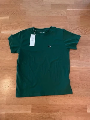 Grön Lacoste Sport t-shirt - Mörkgrön t-shirt från Lacoste Sport. Klassisk modell med rund hals, korta ärmar och den ikoniska krokodilloggan broderad på bröstet. Tillverkad i både polyester och bomull som känns skön mot huden. Perfekt för en clean och sportig look.