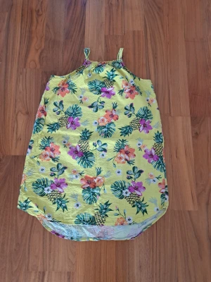 Gul blommig sommarklänning - Söt gul klänning med tropiskt blommönster i grönt, lila och orange. Klänningen har smala axelband och är ärmlös, perfekt för varma dagar. Lätt och luftig modell med rundad nederkant. Perfekt för dig som vill ha en färgglad och somrig look.
