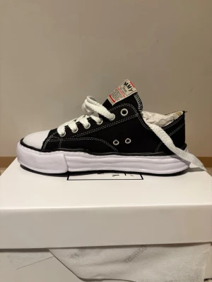 Maison Mihara Yasuhiro (MMY) Peterson  - Unika svarta sneakers från Maison Mihara Yasuhiro med vit, chunky sula och kontrasterande vita sömmar. Skorna har vita skosnören, klassisk låg siluett och detaljerad etikett på plösen. Tillverkade i canvas med gummidetaljer och cool retrovibe.