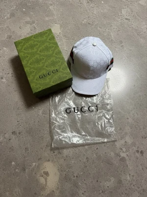Vit keps med monogram - Snygg vit keps med det klassiska GG-monogrammet över hela kepsen. På sidorna finns de ikoniska gröna och röda ränderna. Kepsen har böjd skärm och justerbar rem bak. Kommer med originalkartong och dustbag.