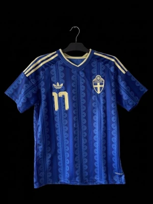 Blå Sverige matchtröja Adidas - Säljer en blå Sverige matchtröja från Adidas med guldiga detaljer och nummer 17. Tröjan har korta ärmar, V-ringning och ett vågigt mönster över hela tyget. På bröstet finns Sveriges landslagsemblem och Adidas-logga. Materialet är troligtvis polyester.