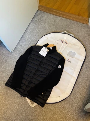 Moncler väst - Snygg svart dunväst från Moncler med stickade svarta ärmar och klassisk Moncler-logga på armen. Västen har quiltad front i glansigt material och dragkedja framtill. Perfekt för lager-på-lager och en stilren look.
