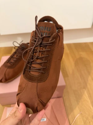 Bruna sneakers från Miu Miu - Unika bruna sneakers från Miu Miu i mjukt läder och satin.Skorna har rundad tå, bruna snören och diskret logga på plösen och tån. Snygg söm längs ovandelen och låg profil gör dem perfekta för en trendig look.