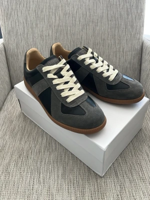 Maison Margiela gats  - Säljer ett par Maison Margiela gats i grå mocka och svart läder med vita snören och klassisk gummisula. En snygg sko med låg profil och diskreta detaljer. Perfekta för dig som gillar stilrena och trendiga sneakers. Tveka inte att höra av er vid frågor🙌🙌