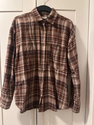 Brun rutig skjorta i bomull - Säljer en brun och beige rutig skjorta med klassisk krage och bröstficka. Skjortan har långa ärmar och knäppning framtill. Perfekt för dig som gillar avslappnad stil och mjuka material.