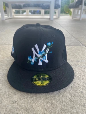 Svart New Era NY keps med blommor - Säljer en svart New Era 59FIFTY keps med NY-logga broderad i vitt och blå blommor på framsidan. Kepsen har en platt skärm och New Era-loggan broderad på sidan. Perfekt för dig som vill sticka ut med en unik och snygg keps. 55cm 
