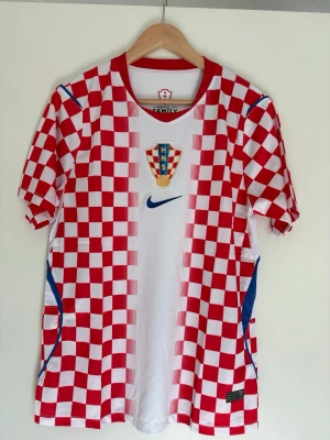 Kroatien fotbollströja Nike röd/vit - Säljer en officiell Kroatien fotbollströja från Nike med klassiskt rödvitt schackrutigt mönster. Tröjan har HNS-emblem på bröstet, blå Nike-logga och detaljer i blått på sidorna. Tillverkad i lätt och ventilerande Dri-FIT material, perfekt för träning eller match.