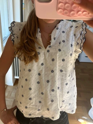Vit blus med volangärm och broderi - Söt vit blus med svarta broderade blommor och volangärmar med svart kant. Blusen har v-ringning och är tillverkad i ett lätt, luftigt tyg som känns perfekt för sommaren. Volangdetaljerna ger en romantisk touch och tyget har diskret struktur.