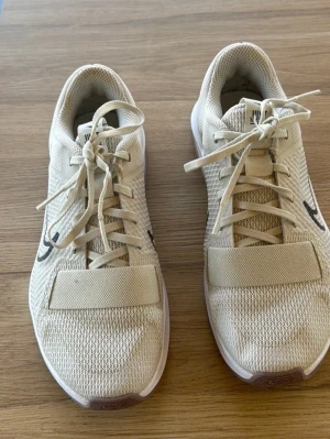 Beige Nike sneakers med mesh och snörning - Säljer ett par beige Nike sneakers med svart swoosh på sidan. Skorna har meshmaterial för bra ventilation, snörning och bred rem över framfoten. Sulan är vit och ger en sportig look. Perfekta för dig som gillar stilrena och bekväma sneakers.