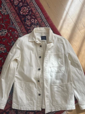 Vit overshirt från ARKET i bomull - Stilren vit overshirt från mango i  bomull. Modellen har tre stora fickor, klassisk krage och svarta knappar framtill. Perfekt lager-på-lager-plagg med avslappnad passform och tidlös look.