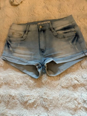 Ljusblå denimshorts med slitningar - Säljer ett par ljusblå denimshorts med snygga slitningar och uppvikta benslut. Klassisk femficksmodell med knapp och dragkedja framtill. Perfekta för varma dagar och ger en avslappnad look.