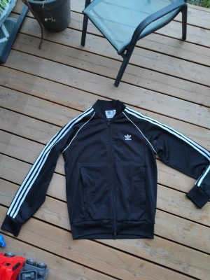 Svart Adidas track jacket - Klassisk svart Adidas track jacket med vita ränder längs ärmarna och vit Adidas-logga på bröstet. Dragkedja framtill, ribbade muddar och krage. Tillverkad i ett mjukt och stretchigt material som passar perfekt till träning eller streetwear.