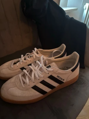 Adidas Spezial vita sneakers - Säljer ett par Adidas Spezial sneakers i vitt mocka med klassiska svarta ränder på sidorna och brun gummisula. Skorna har låg profil och vita skosnören. Perfekta för dig som gillar retrostil och streetwear.