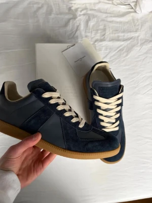 Mörkblå sneakers från Maison Margiela - Snygga mörkblå sneakers från Maison Margiela med ovandel i mocka och läder, vita skosnören och klassisk gummisula. Ikonisk design med retrovibbar och diskreta detaljer. Perfekta för dig som gillar stilrena och tidlösa sneakers.