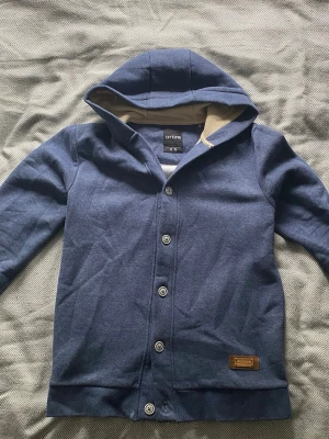 Blå hoodie med knappar från Ombre - Snygg blå hoodie från Ombre i storlek M med knappar framtill och beige foder i huvan. Hoodien har långa ärmar och en liten läderdetalj på nederkanten. Perfekt för dig som gillar stilren design och mjuka material.