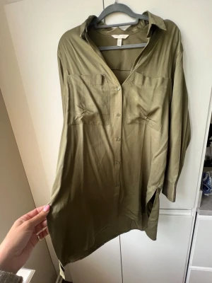 Olivgrön storskjorta från H&M - Säljer en olivgrön storskjorta från H&M med klassisk krage, knappar framtill och två bröstfickor. Skjortan har lång ärm och är tillverkad i ett mjukt, lätt material som ger en avslappnad känsla. Perfekt för en chill och stilren look. 