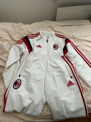 Vit AC Milan Adidas träningsdress - Säljer en vit träningsdress från Adidas med AC Milan-logga på både jacka och byxor. Dressen har röda och svarta detaljer, dragkedjor på jackan och byxorna samt klassiska Adidas-ränder längs ärmar och ben. Perfekt för fotbollsträning eller chill.