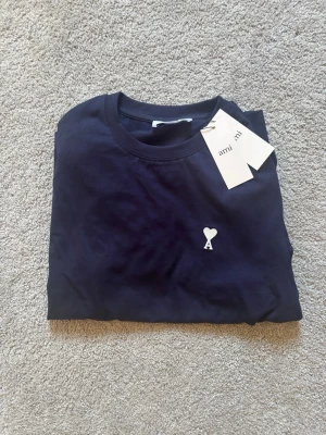Mörkblå t-shirt från Ami Paris - Snygg mörkblå t-shirt från Ami Paris med klassisk passform och rund hals. T-shirten har ett litet vitt hjärta med bokstaven A broderat på bröstet. Tillverkad i mjuk bomull som känns skön mot huden. Perfekt till jeans eller shorts.
