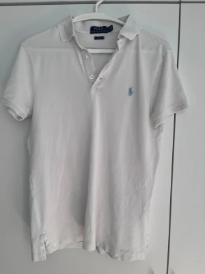Vit pikétröja från Polo Ralph Lauren - Klassisk vit pikétröja från Polo Ralph Lauren med korta ärmar och blå broderad logga på bröstet. Tröjan har krage, knappar framtill och är tillverkad i mjuk bomull. Perfekt till en avslappnad och stilren look.