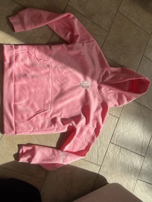Rosa hoodie med broderad logga - Vid mer bilder på dem alla broderingarna är det bara att skriva. Snygg rosa hoodie med huva och magficka. Broderad vit logga på bröstet och diskret textdetalj på ena ärmen. Tillverkad i mjukt material som känns skönt mot huden. Perfekt för en avslappnad och trendig look. 