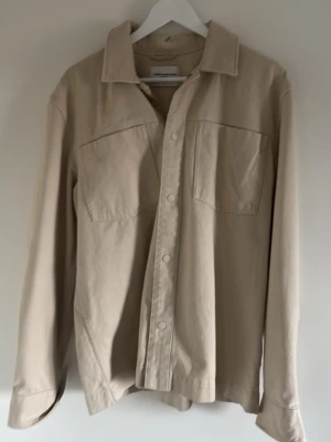 Beige skjorta från These Glory Days - Stilren beige overshirt från These Glory Days i klassisk modell. Overshirten har långa ärmar med knappar vid ärmsluten och är tillverkad i ett mjukt bomullstyg. Perfekt för en clean och avslappnad look.