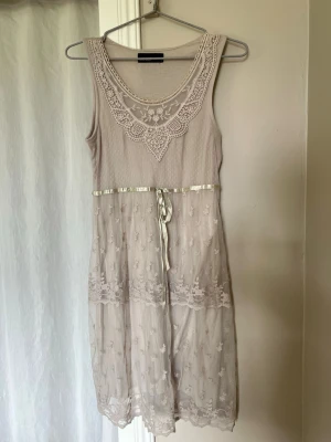 Beige spetsklänning från Vero Moda - Supersöt beige klänning från Vero Moda med spetsdetaljer och broderad halsringning. Klänningen är ärmlös och har ett band i midjan samt transparent spets med blommönster på nederdelen. Perfekt för dig som gillar romantiska och feminina plagg.