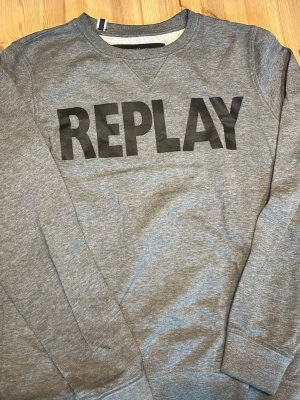 Grå hoodie från Replay - Snygg grå hoodie från Replay med stort svart logotryck på bröstet. Skit skön att ha på sig i jätte najs matrial. Och snygg färg och i helt nytt skick. I storlek L men sitter bra på mig som har M, kan skicka bild ifall det behövs!