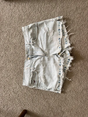Ljusa denimshorts från Hollister - Säljer ett par ljusa denimshorts från Hollister med slitna detaljer och fransig kant. Shortsen har broderade blommor längs bensluten och klassiska fem fickor. Perfekta för varma dagar och ger en avslappnad vibe.