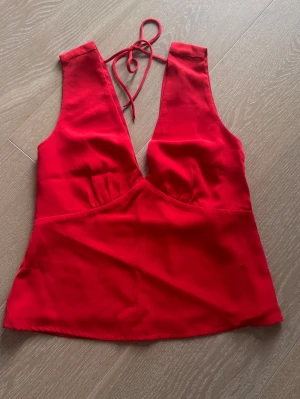 Röd topp blus med djup v-ringning i stl XS helt ny - Snygg röd linnetopp med djup v-ringning och knyt i nacken. Toppen är ärmlös och har en figurnära passform som framhäver siluetten. Perfekt för dig som vill sticka ut med en färgstark och trendig look.