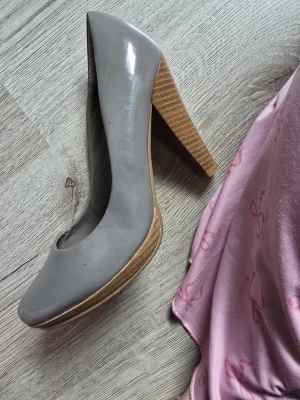 Grå pumps med träklack - Snygga grå pumps från Shoes by Marco med rundad tå och hög träklack. Skorna har glansig finish och insidan är fodrad med leopardmönster för en cool kontrast. Perfekta för dig som vill ha en stilren men ändå unik look.