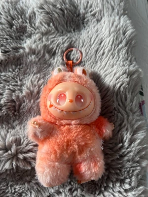 Söt orange fluffig nyckelring - Unik nyckelring med en orange fluffig figur som har ett plastansikte med stora ögon och ett brett leende. Figuren har små öron och är tillverkad av mjukt plyschmaterial med plastdetaljer. Perfekt som accessoar till väskan eller nycklar.