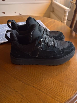UGG lowmel  - Snygga svarta sneakers från UGG med robust sula och detaljer i mocka och textil. Skorna har tjocka, mönstrade skosnören och en vadderad plös med UGG-logga. Perfekta för dig som gillar en chunky och sportig look.