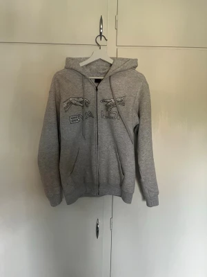 Bvaldi hoodie  - Grå hoodie med dragkedja från Bvalid. Har två fickor framtill, huva med dragkedja och tryckt logga med fågelmotiv på bröstet. Perfekt för en avslappnad och stilren look. Mjuk och skön bomullsblandning. Kostar 1299kr ny. Den har Inte tappat några pärlor eller något sån, det är bara att höra av er om ni undrar något