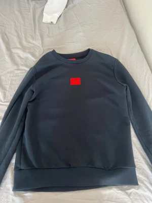 Mörkblå HUGO sweatshirt  - Snygg mörkblå sweatshirt från HUGO med en ikonisk röd logotyp på bröstet. Tröjan har rund halsringning, ribbade muddar och är tillverkad i mjukt material som känns skönt mot huden. Perfekt för en stilren och avslappnad look.