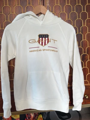 Vit hoodie från Gant med tryck - Snygg vit hoodie från Gant med broderad logga och texten 'American Sportswear' på bröstet. Klassisk känguruficka framtill och huva. Perfekt för en avslappnad och sportig stil.