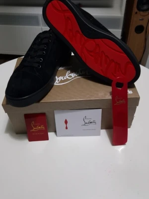Svarta Louboutin sneakers med röd sula - Snygga svarta sneakers från Christian Louboutin med ikonisk röd sula och diskret logga. Skorna har lågt skaft, svarta skosnören och är tillverkade i mocka. Perfekta för dig som vill sticka ut med exklusiv stil.