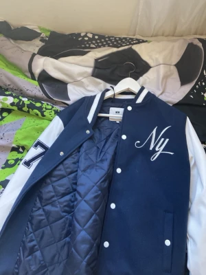 Blå bomberjacka från New Garments - Snygg blå bomberjacka med vita ärmar från New Garments. Jackan har broderad 'Ny' på bröstet, quiltat foder och tryckknappar framtill. Ärmarna är vita med en blå '7' på vänster arm. Klassisk college-stil med ribbade muddar och krage.