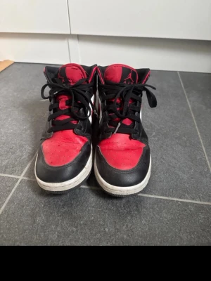 Air Jordan 1 Mid svart/röd/vit - Säljer ett par Air Jordan 1 Mid sneakers i svart, rött och vitt läder med klassisk Nike Swoosh på sidan. Skorna har svarta skosnören, perforerad tåbox och ikoniska Jordan Wings-loggan vid ankeln.  Sulorna är bytt till ett nytt par för en skönare och bättre passform. 