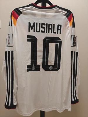 Tysklands spelar version fotbollströja Musiala #10 - Vit långärmad Tyskland fotbollströja från Adidas med Musiala och nummer 10 på ryggen. Tröjan har svarta, röda och gula detaljer, officiella FIFA World Cup-märken på ärmarna och klassiska svarta ränder längs ärmarna. Perfekt för fotbollsfans!