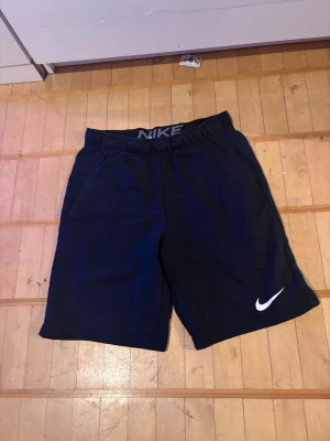 Svarta Nike shorts med logga - Säljer ett par svarta shorts från Nike med elastisk midja och vit Nike-logga på vänster ben. Shortsen har en enkel och sportig design, perfekta för träning eller chill. Tillverkade i mjukt material för skön passform.